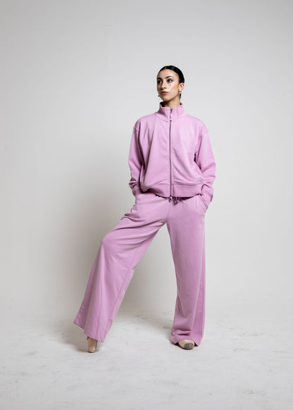 A model wearing a matching lavender pink tracksuit with a zip-up jacket and wide-leg pants, standing against a plain light gray studio background.