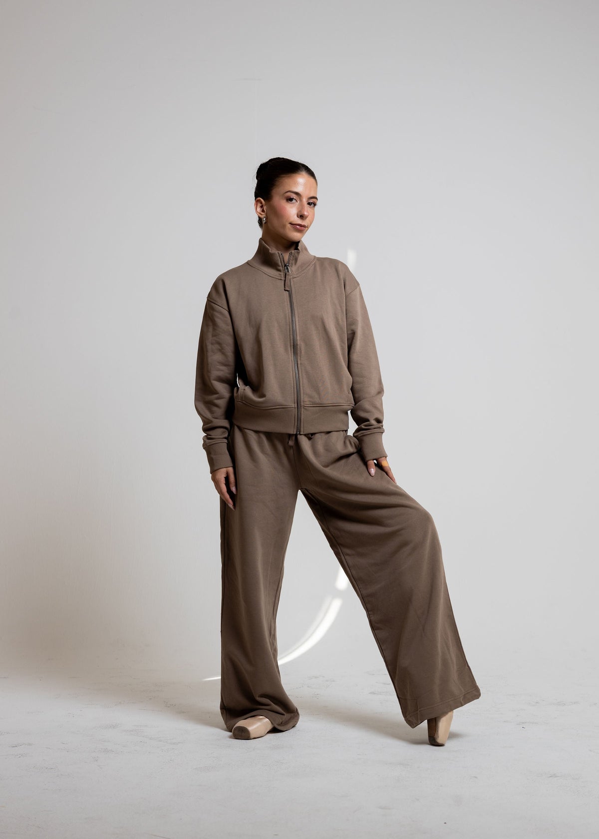 A woman modeling a matching taupe-brown two-piece tracksuit with a zip-up jacket and wide-leg pants, posed against a neutral light gray studio background.
