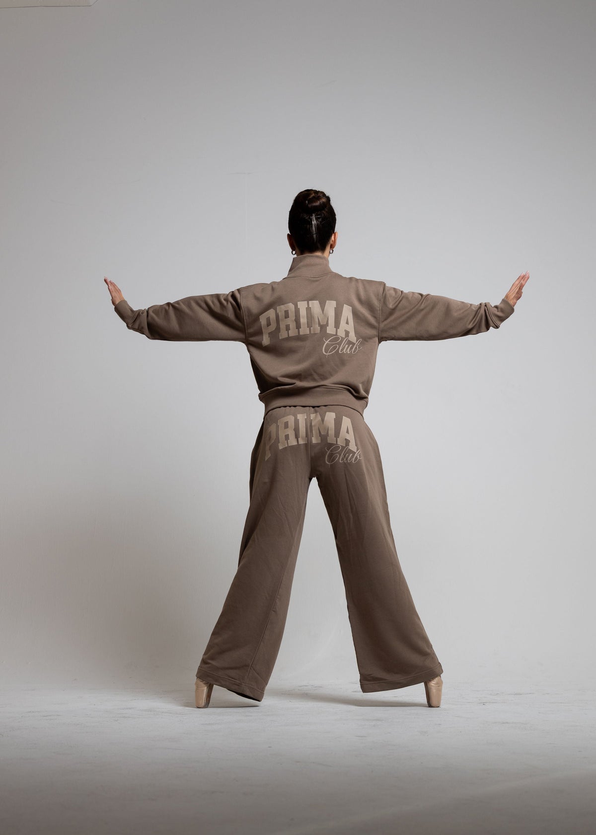 A model is shown from behind wearing a matching taupe-brown tracksuit with 'PRIMA CLUB' printed in beige on the back of the jacket and pants, posed with arms outstretched against a neutral gray studio background.