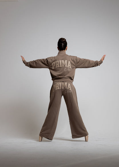 A model is shown from behind wearing a matching taupe-brown tracksuit with 'PRIMA CLUB' printed in beige on the back of the jacket and pants, posed with arms outstretched against a neutral gray studio background.