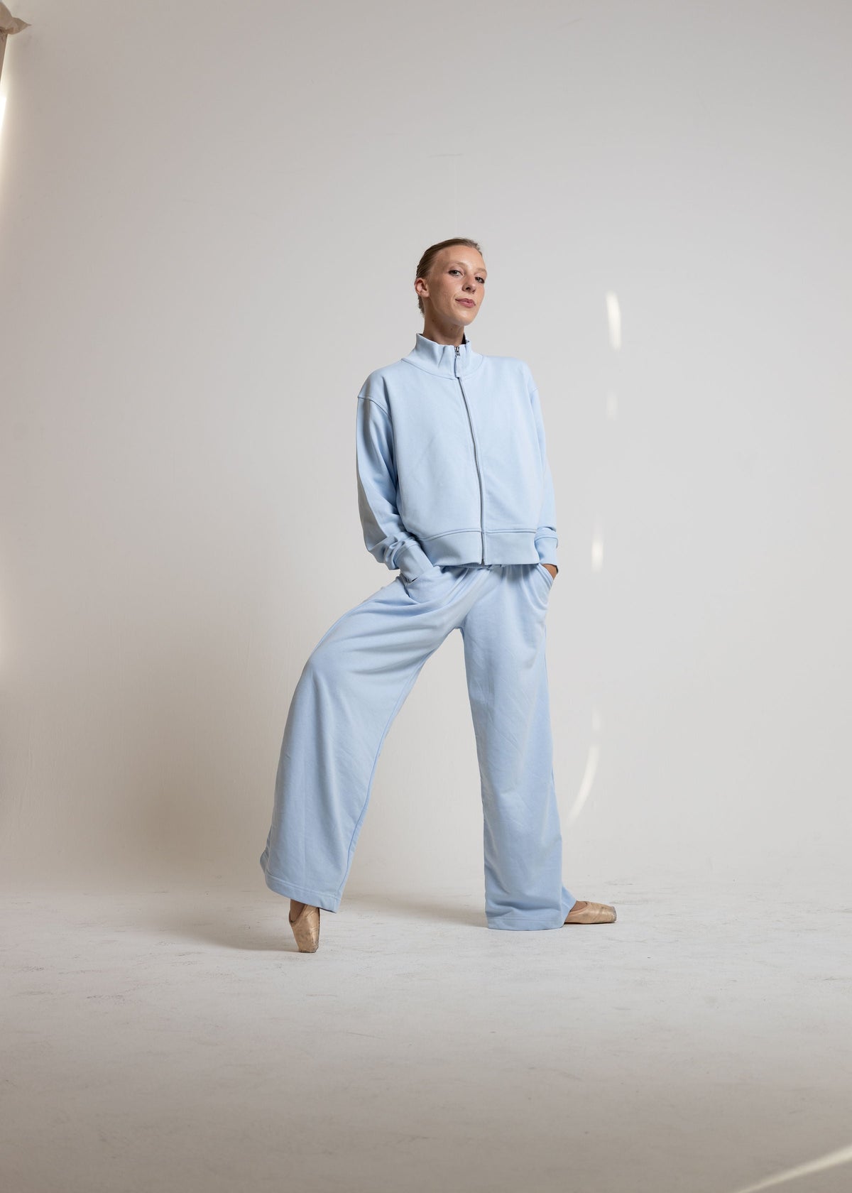 A model wearing a light blue two-piece tracksuit consisting of a cropped zip-up jacket and matching wide-leg trousers, standing in a minimalist studio setting.
