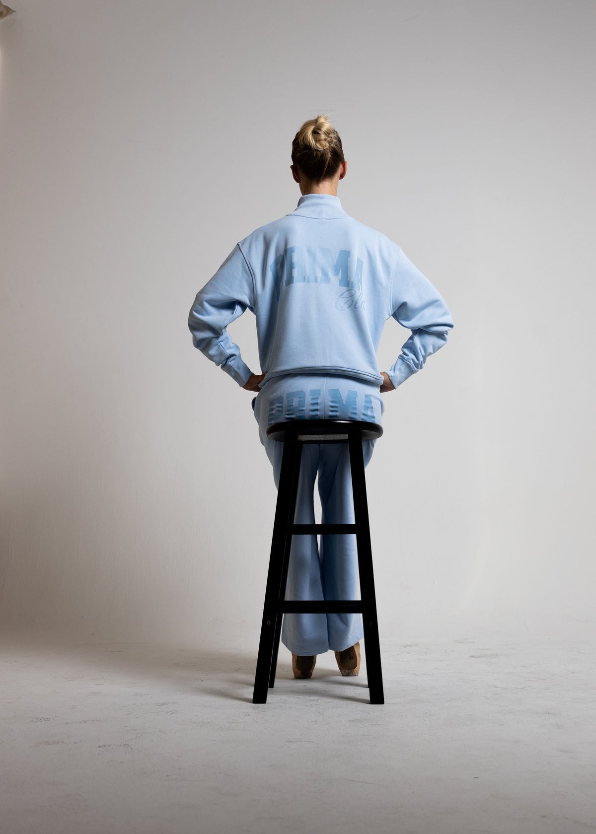 A model seen from behind wearing a light blue sweatshirt and matching sweatpants, sitting on a black stool in a minimalist studio setting.