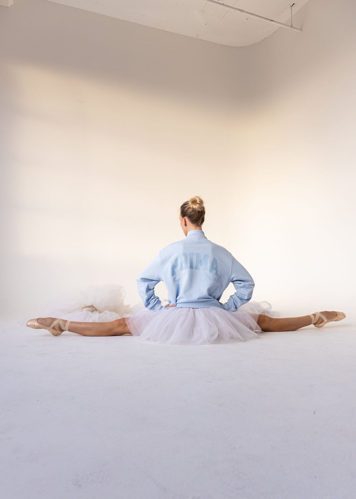 A woman in a ballet tutu and light blue denim jacket sits on the floor in a split position, viewed from behind in a bright, minimalist studio.