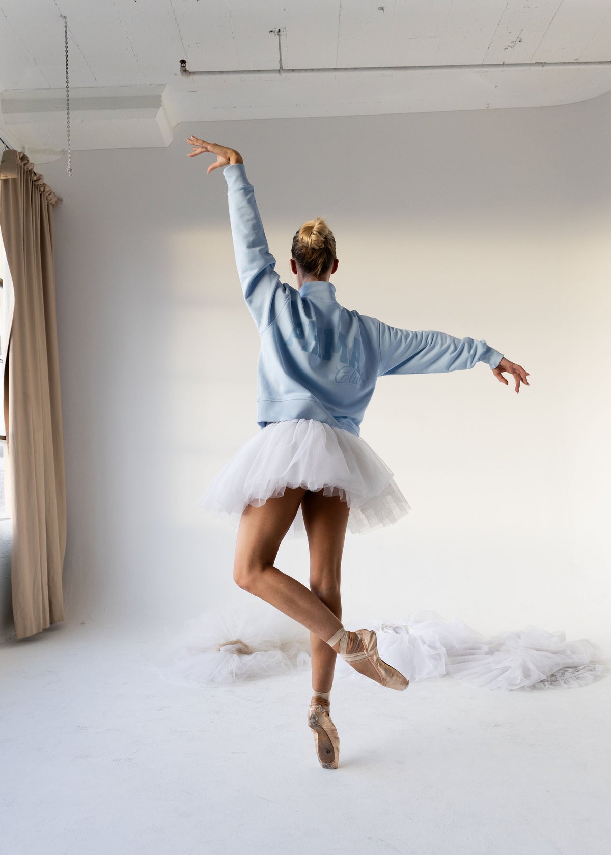 A woman in a light blue sweatshirt and white tutu skirt dances in a bright white studio, shown from behind with one arm raised and the other extended side to side.