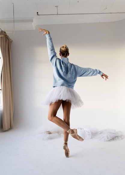 A woman in a light blue sweatshirt and white tutu skirt dances in a bright white studio, shown from behind with one arm raised and the other extended side to side.
